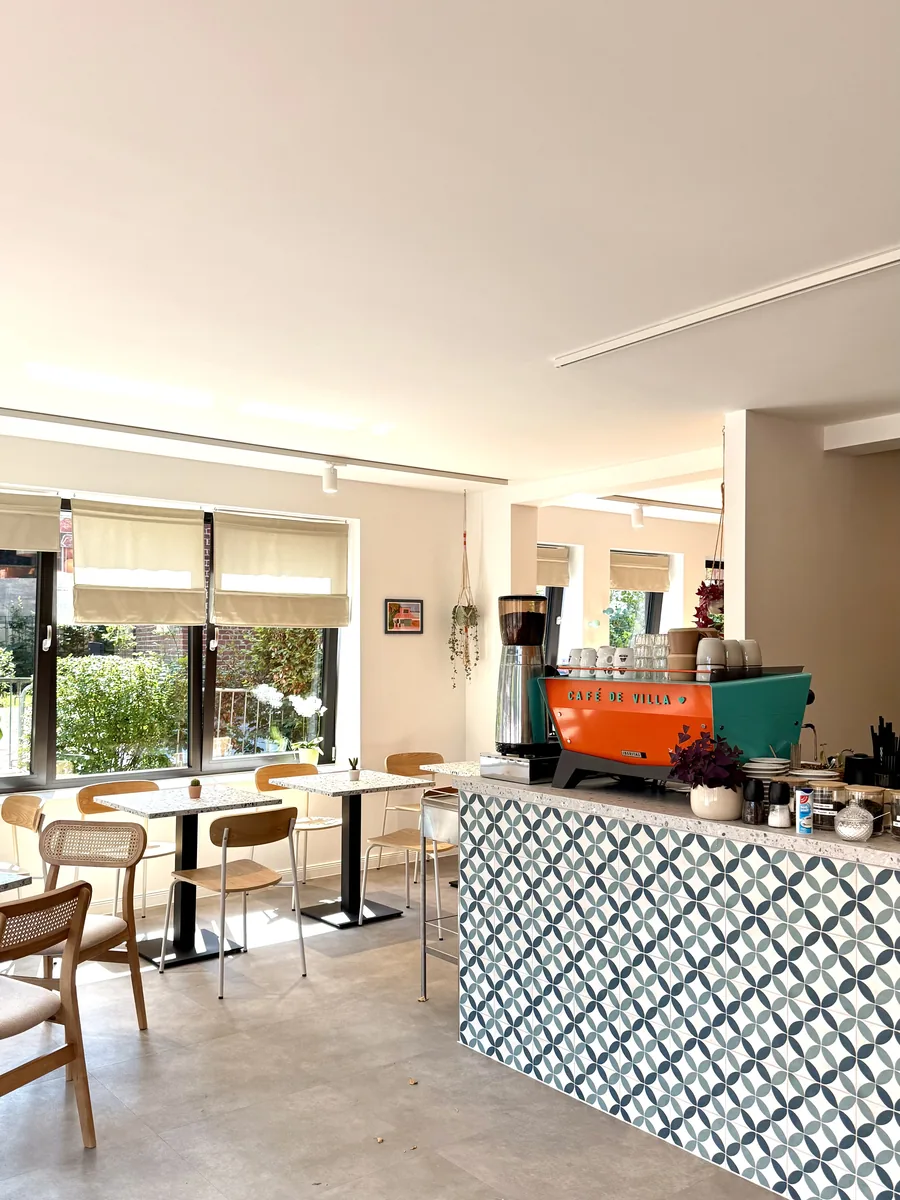 Heller Gastraum des Café de Villa mit markanter oranger Espressomaschine an gemusterter Theke
