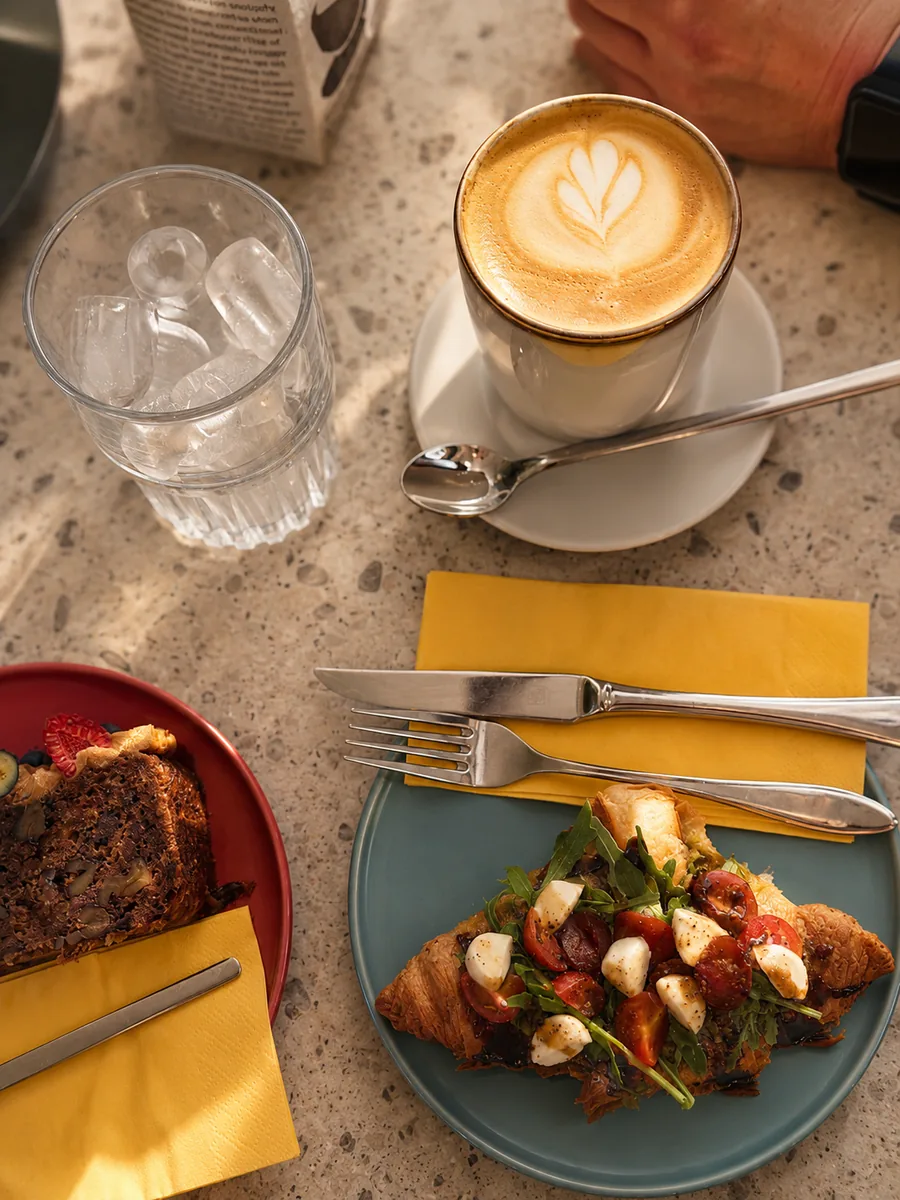 Cappuccino mit Latte Art, belegtes Croissant mit Mozzarella und Tomaten sowie Kuchen im Cafe de Villa